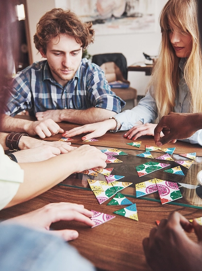 Een diverse groep van vijf mensen concentreert zich intens op het in elkaar zetten van kleurrijke driehoekige puzzelstukken op een houten tafel, wat samenwerking en concentratie uitstraalt.