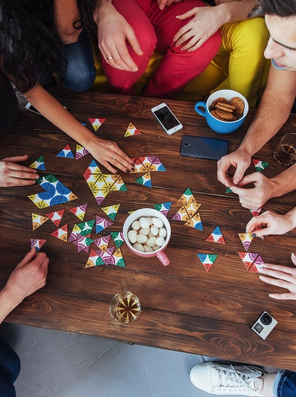 Groep vrienden geniet van een spel Jenga aan een buitentafel. Een deelnemer bedekt zijn gezicht van het lachen, terwijl anderen glimlachen en klappen. Ontspannen, vrolijke sfeer.